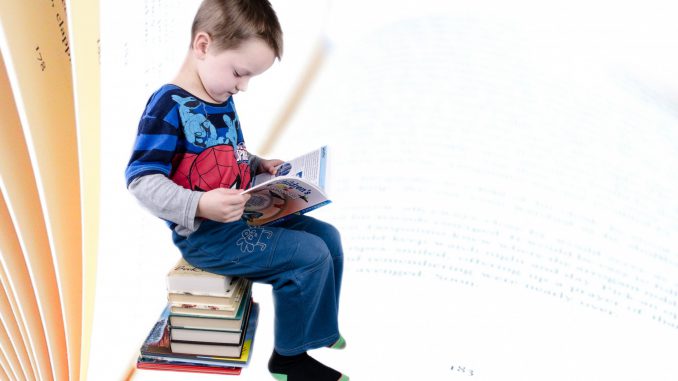 Na zdjęciu chłopiec z ksiązką siedzący na stosie książek