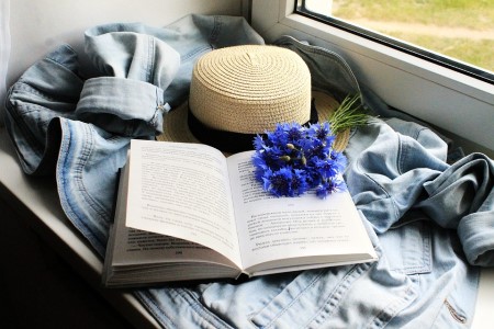 Na zdjęciu parapet okna w pociągu a na nim otwarta książka kapelusz słomkowy i bukiet chabrów