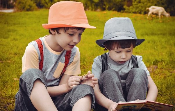 Na zdjaciu dwaj chłopcy w kapeluszach siedzą na łąće. Przed jednym otwarta książka.